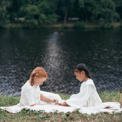 two girls in white dresses sitting on riverbank